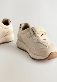 Baskets de sport beige avec une tige synthétique lisse, des accents texturés, un système de fermeture à lacets et Velcro, et une semelle en caoutchouc avec des motifs de adhérence.