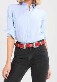 Camisa de botones de color azul claro con rayas verticales blancas, mangas arremangadas, combinada con jeans negros y un cinturón rojo con detalles plateados.