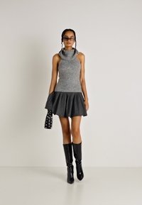 Maglione senza maniche grigio con colletto alto abbinato a una gonna nera plissettata, stivali neri fino al ginocchio e una borsa nera testurizzata con borchie.