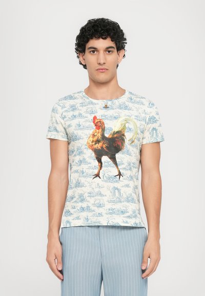 Jonge man met krullend haar die een kortemouwen wit shirt draagt met een grote kleurrijke haan en blauwe gebloemde shorts.
