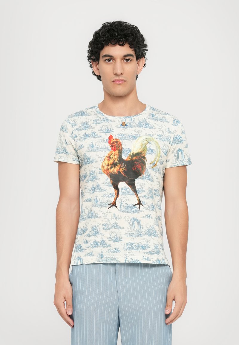 Camiseta de manga corta en color crema con un gráfico colorido de un gallo, rodeado de un patrón repetido en azul que representa escenas pastorales.