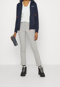 Veste à capuche bleu marine avec des accents orange, débardeur bleu clair, pantalon gris et bottes de randonnée noires, tenant une bouteille d'eau gris foncé.
