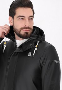 Juodas vandeniui atsparus švarkas su kapišonu, raišteliais, metaliniu užtrauktuku ir logotipu ant krūtinės. Lygi tekstūra, minimalistinis dizainas.