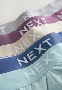 Pile de bandes de tissu en gris, violet, beige, bleu et bleu clair, chacune avec le mot "NEXT" écrit en lettres majuscules blanches.
