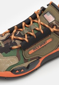 Camouflage tenisky s kombinací zelené, hnědé a černé textury. Mají oranžové šněrování a akcenty, gumovou podrážku a branding 'POLO SPORT'.