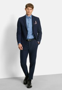 Navy suit with a fitted design, light blue shirt, and patterned pocket square. Black belt and polished shoes complete the outfit.