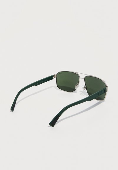 Ορθογώνια γυαλιά ηλίου με πράσινους φακούς, ασημένιο μεταλλικό σκελετό και σκοτεινά πράσινα μπράτσα, τοποθετημένα επίπεδα σε λευκή επιφάνεια.