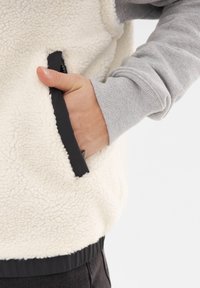 Krämfärgad fleeceväst med svart kant, med en sidoficka och en grå långärmad skjorta under. Mjuk textur och avslappnad design.