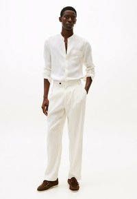 Camisa de lino blanca con un frente abotonado y bolsillo, combinada con pantalones crema a rayas y zapatos marrones.