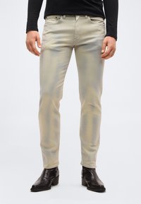 Άνδρας που φοράει στενά μπεζ τζιν, μαύρο μακρυμάνικο πουκάμισο και μαύρα δερμάτινα μποτάκια μέχρι τον αστράγαλο, στέκεται σε απλό ανοιχτόχρωμο φόντο.