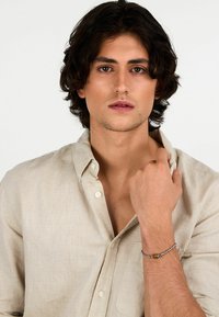 Braccialetto d'argento con un design texture e una perla marrone, indossato su un polso, su uno sfondo neutro. Camicia di lino casual beige.