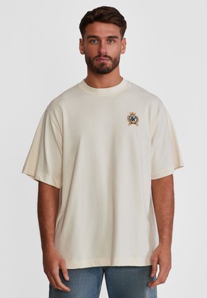 Mann med kort mørkt hår og skjegg som har på seg en stor kremfarget t-skjorte med lite brodert emblem på venstre bryst og blå jeans.