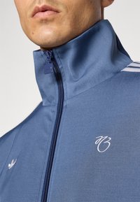 Jachetă sport albastră cu guler înalt, fermoar complet și logo brodat alb. Are trei dungi albe pe un umăr. Textură de material neted.