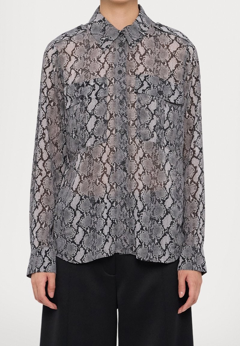Chemise à manches longues boutonnée avec un motif serpent gris et noir, deux poches sur la poitrine, portée avec un pantalon noir.