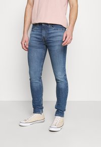 Hombre con jeans ajustados azul, camiseta rosa clara y zapatillas altas beige, de pie sobre un suelo gris frente a una pared blanca.