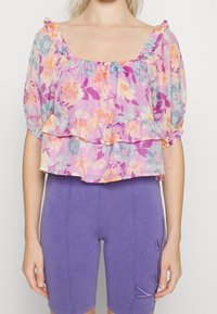 Top court à imprimé floral en tissu doux et léger avec un col et des manches volantés. Présente des fleurs vibrantes de couleur rose, violet, orange et bleu.