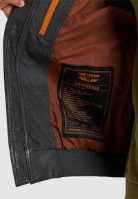 Zwarte leren jassen interieur met een oranje rits en bruine voering. Bevat een productlabel met specificaties en details over de zakken en manchetten.