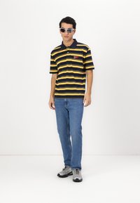 Polo skjorte med marineblå og gule horisontale striber, korte ærmer og et lommemonogram. Båret med blå jeans og grå sportsko.