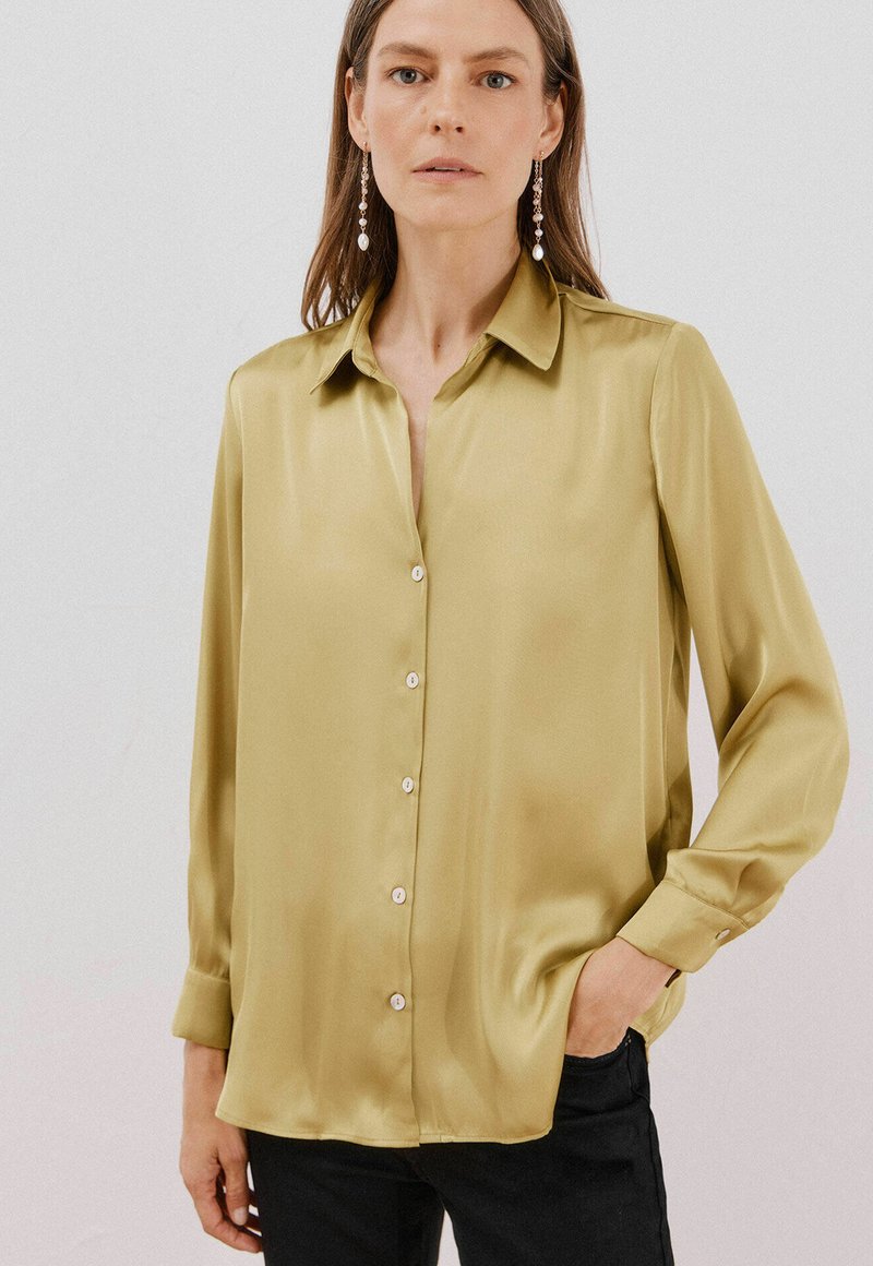 Blusa in satin a maniche lunghe di colore verde oliva con colletto a punta, chiusura a bottoni sul davanti e dettagli a bottoni. Tessuto liscio e vestibilità comoda.