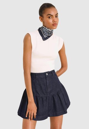 Jonge vrouw draagt een witte mouwloze top, donkere uitlopende spijkerrok en een zwarte patroon halsdoek, poserend tegen een effen achtergrond.