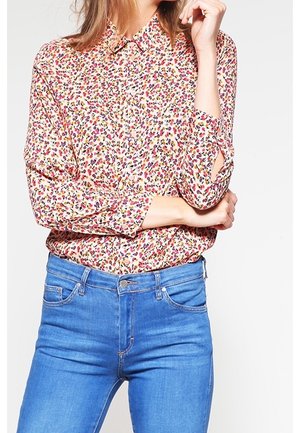 Woman wearing a long-sleeve button-up shirt with a multicolored floral print tucked into high-waisted blue jeans.
