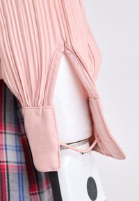Light pink pleated blouse with a rounded collar and open cuffs, featuring a textured finish and an accentuated hemline.