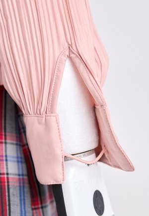 Light pink pleated blouse with a rounded collar and open cuffs, featuring a textured finish and an accentuated hemline.