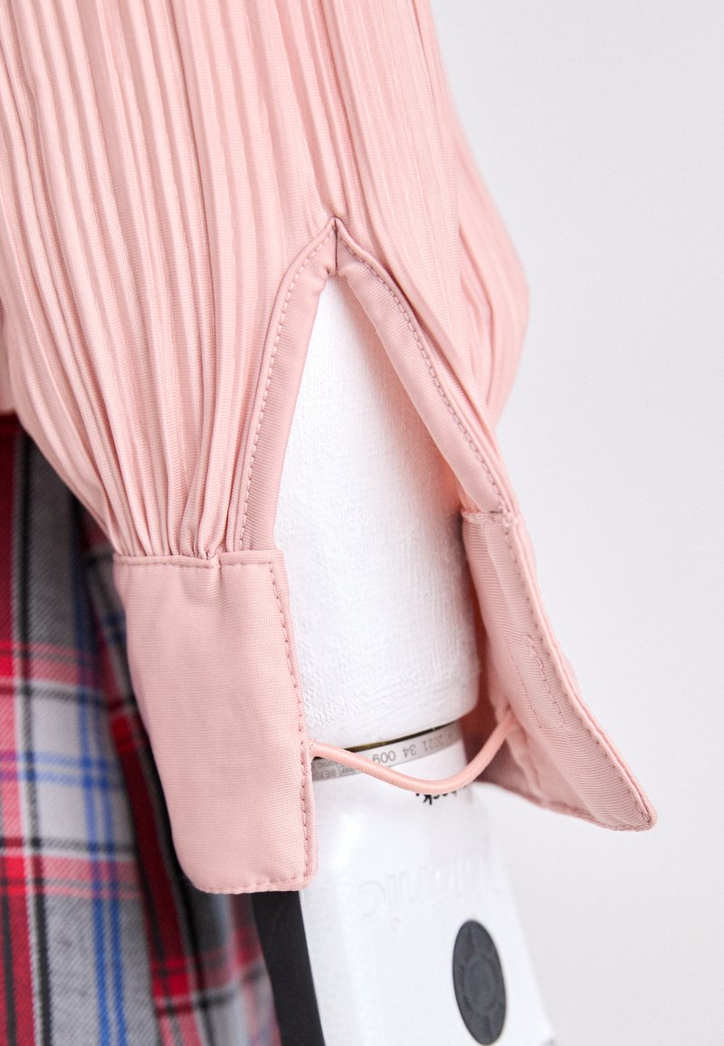 Light pink pleated blouse with a rounded collar and open cuffs, featuring a textured finish and an accentuated hemline.