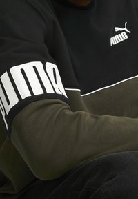 Sudadera negra y verde oliva con el logo blanco de Puma. Presenta una sección superior negra, una inferior verde y una gran marca blanca en la manga.