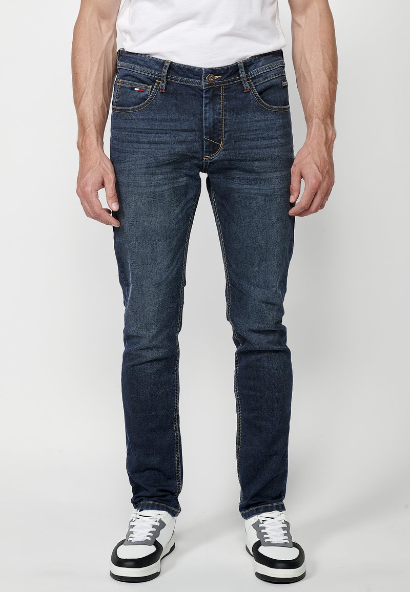 Slim fit tmavé denimové džíny s kontrastním šitím, klasickým pěti kapsovým provedením a zipem, v kombinaci s bílými a černými teniskami.