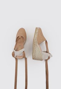 Zapatos de espadrilles de ante marrón con una correa de lona trenzada para el tobillo y lazos de tela marrón. Taco de cuña envuelto en cuerda gruesa. Textura suave.