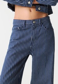 Section médiane vêtue d'un jean bleu et d'une veste à fines rayures blanches verticales, laissant voir le ventre et le nombril nus.