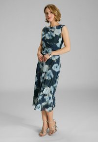 Blumenbedrucktes, ärmelloses Kleid in dunklem Petrol, mit blauen und grünen Mustern und einer fließenden Silhouette. Abgerundet mit silbernen Riemchensandalen.
