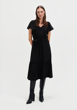Czarna midi sukienka z dekoltem w kształcie litery V, krótkimi falbaniastymi rękawami i związanym w talii pasem. Posiada falbaniaste spódnice, zestawiona z czarnymi kozakami za kolano.
