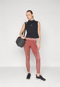 Haut sans manches noir, leggings ajustés couleur rouille avec des motifs géométriques, et baskets noires. Tient un sac de sport gris foncé.