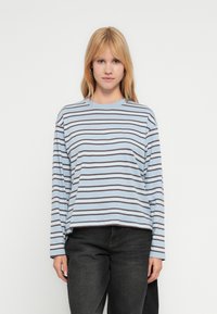 Ανοιχτό μπλε T-shirt με μακριά μανίκια και οριζόντιες σκούρες καφέ ρίγες, στρογγυλή λαιμόκοψη και άνετη εφαρμογή, συνδυασμένο με σκούρο τζιν παντελόνι.