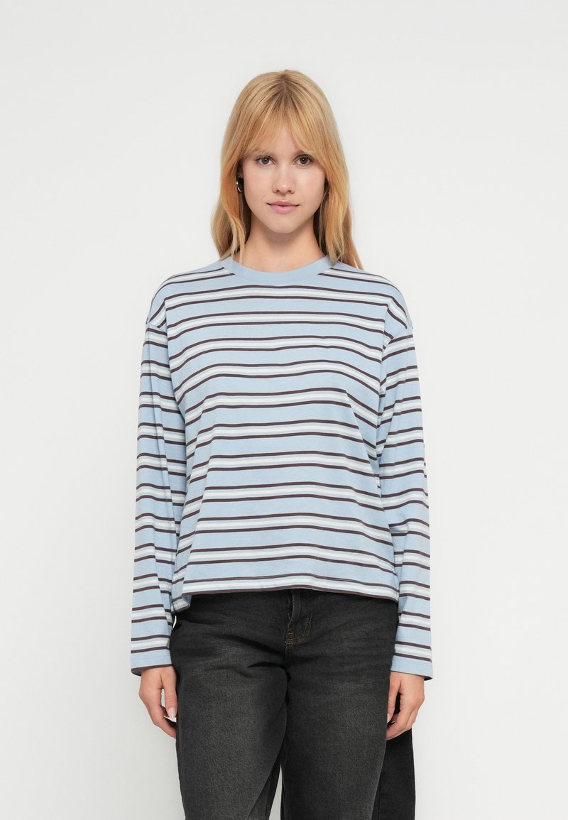 Ανοιχτό μπλε T-shirt με μακριά μανίκια και οριζόντιες σκούρες καφέ ρίγες, στρογγυλή λαιμόκοψη και άνετη εφαρμογή, συνδυασμένο με σκούρο τζιν παντελόνι.