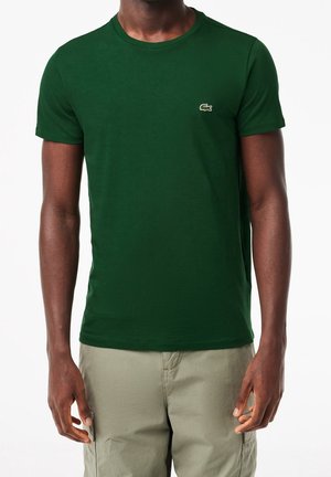 Mann trägt dunkelgrünes T-Shirt mit kurzem Arm und Rundhalsausschnitt, mit kleinem Krokodil-Logo auf der Brust und hellbeige Hose.