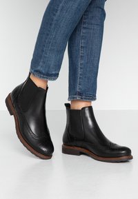 Stivali Chelsea neri in pelle con pannelli elastici laterali, dettagli brogue e suola cucita marrone, indossati con jeans blu.
