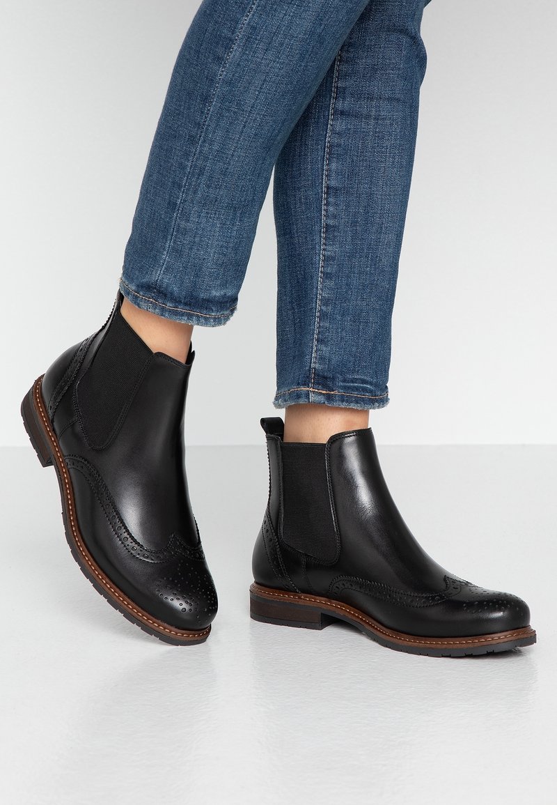 Stivali Chelsea neri in pelle con pannelli elastici laterali, dettagli brogue e suola cucita marrone, indossati con jeans blu.