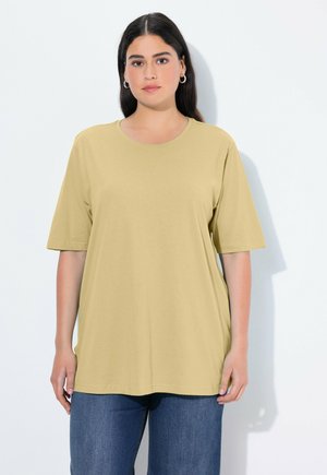 Frau mit langem dunklem Haar trägt ein lockeres hellgelbes kurzärmeliges Hemd und blaue Jeans, steht vor einem schlichten hellen Hintergrund.