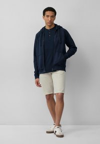 Felpa con zip blu navy, con cordini, abbinata a un polo blu scuro e pantaloni corti beige chiaro. Sneakers bianche con dettagli marroni completano l'outfit.