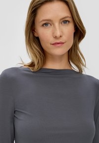 Vrouw met blond haar en blauwe ogen, gekleed in een grijze bootneck top, kijkt recht in de camera met een neutrale uitdrukking.