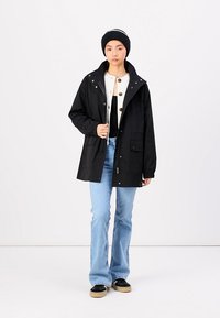 Veste imperméable noire avec capuche, boutons à l'avant et poches. Associée à un jean évasé bleu clair et des chaussures noires. Bonnet avec une rayure blanche.