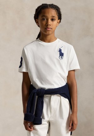 Une jeune personne se tient face à l’objectif, vêtue d’un T-shirt blanc à logo et d’un pantalon, avec un pull bleu marine noué autour de la taille.