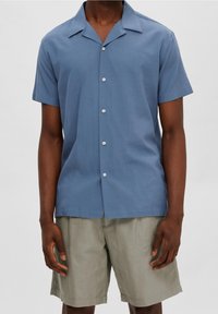 Kortärmad skjorta med krage i ljusblått tyg med mjuk textur och sex vita knappar längs framsidan. Matchad med ljusbeige shorts.