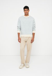 Férfi áll egy fehér háttér előtt, világoskék és fehér mintás pulóverben, bézs nadrágban és fehér sportcipőben.