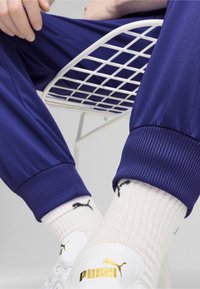 Purple athletic pants with ribbed cuffs, paired with cream socks featuring black logos and white sneakers displaying gold branding.