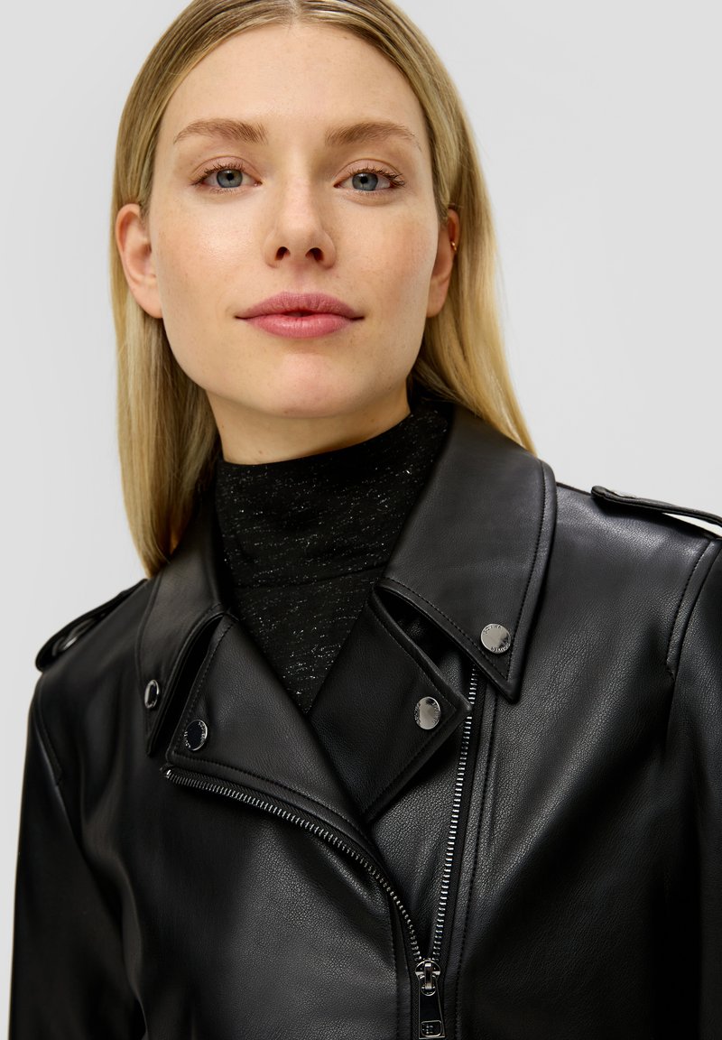 Schwarze Leder-Motorradjacke mit silbernen Beschlägen, Reißverschluss und Schulterklappen. Über einem schwarzen, strukturierten Rollkragenpullover getragen.