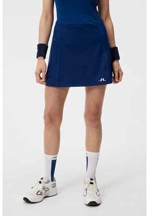 Žena v tmavě modré sportovní sukni s malým bílým logem, bílých a modrých ponožkách, bílých teniskách a tmavě modrých náramcích na zápěstí.